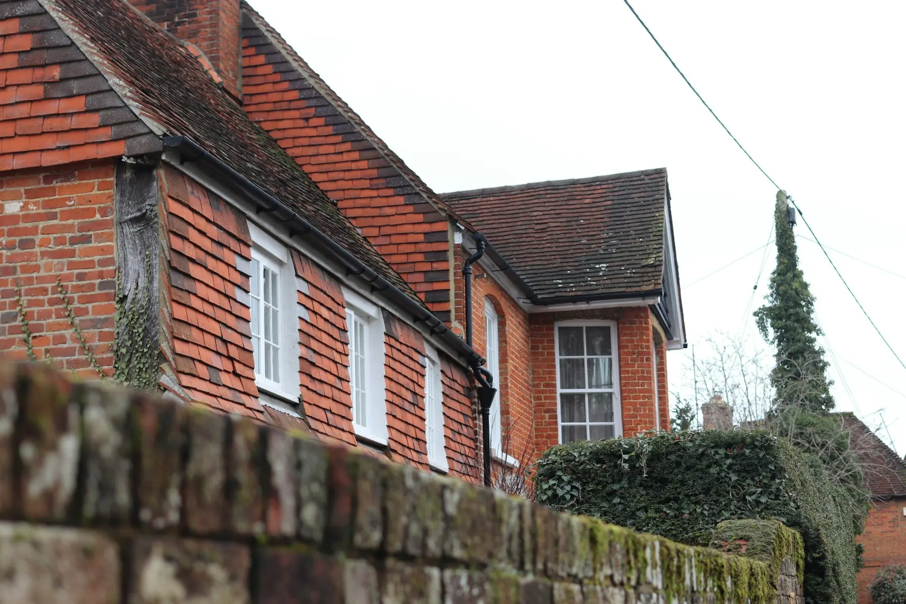 A brick and tile-hung house with white windows and a tiled roof, seen over a garden wall